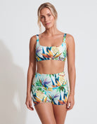 A woman modeling a matching tropical print bikini set against a plain white studio background. The set includes a scoop-neck bikini top and high-waisted bikini shorts with a colorful leaf and bird-of-paradise pattern.
