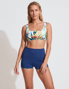 A woman modeling a two-piece swimwear set consisting of a tropical print sports bra and high-waisted navy blue shorts, set against a plain light gray studio background.