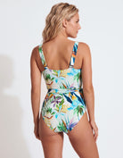 A woman modeling a one-piece swimsuit from the back, showing a tropical floral print with blue, green, orange, and purple leaves on a white background.