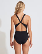 A woman shown from the back wearing a black one-piece swimsuit with crisscross shoulder straps and an open back, photographed against a light gray studio background.