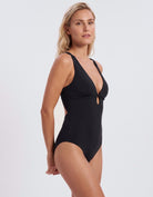 A blonde woman modeling a black one-piece swimsuit with a deep V-neckline, side cutouts, and a small front keyhole detail, photographed against a light gray studio background.