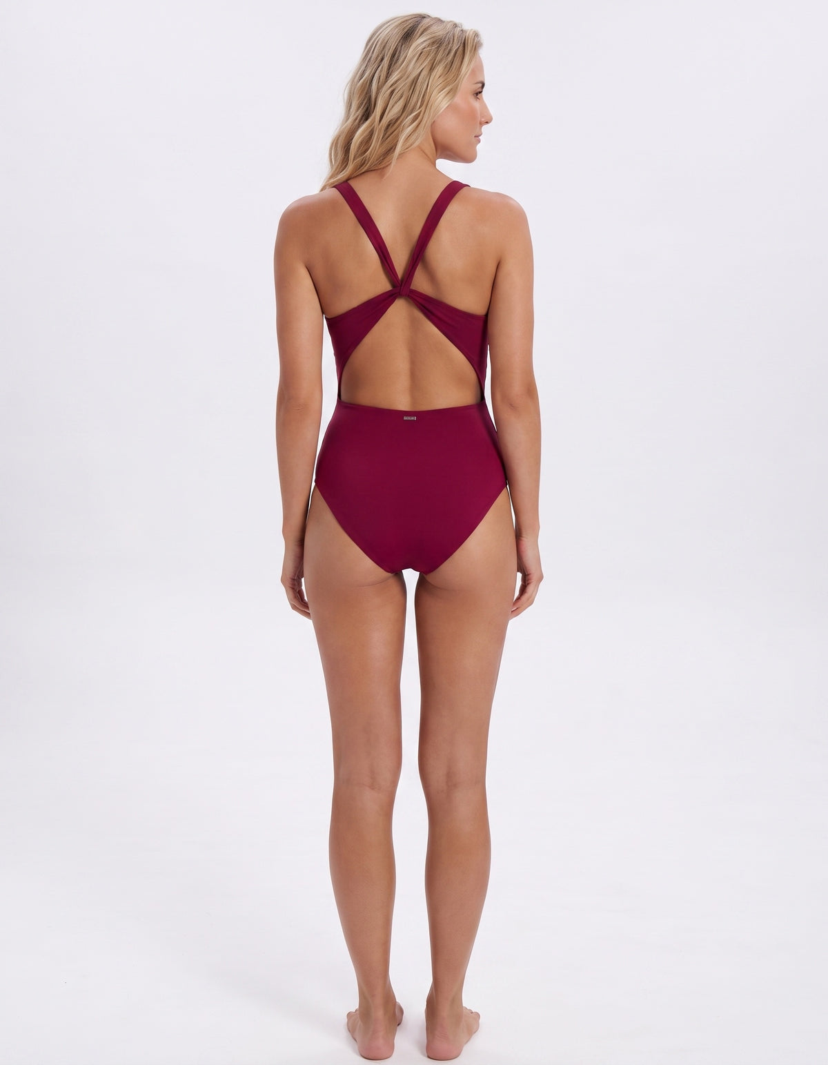 A woman shown from the back wearing a one-piece swimsuit in deep burgundy, featuring a high-cut bottom and an open crisscross back design, set against a plain white studio background.