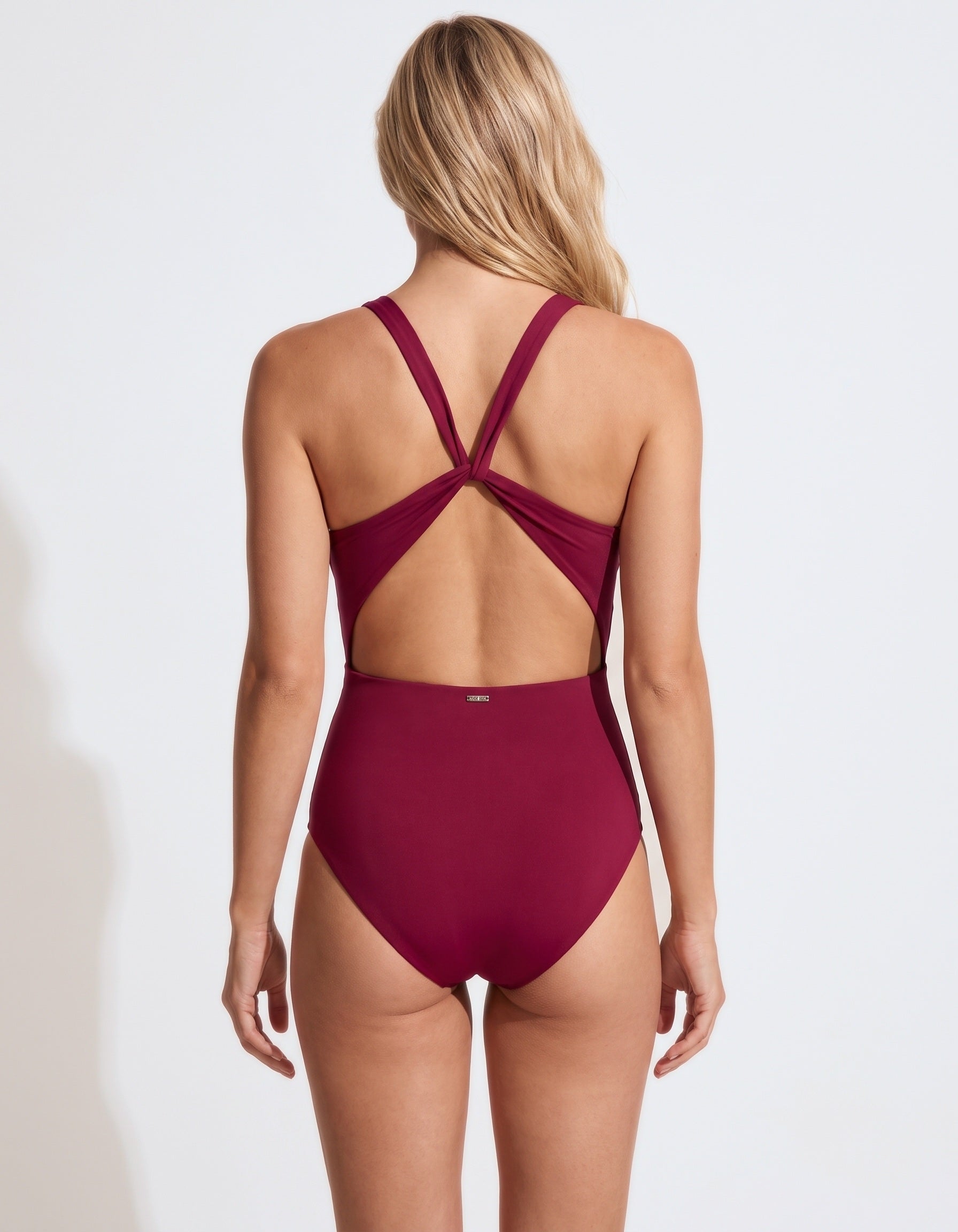 A woman viewed from the back wearing a high-waisted one-piece swimsuit in deep burgundy. The swimsuit features crisscrossing back straps and a small gold logo on the center back waistband.