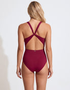 A woman viewed from the back wearing a high-waisted one-piece swimsuit in deep burgundy. The swimsuit features crisscrossing back straps and a small gold logo on the center back waistband.