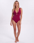 A woman modeling a deep V-neck one-piece swimsuit in a rich burgundy color, standing against a seamless white studio background.