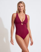 A woman modeling a deep V-neck one-piece swimsuit in burgundy with a small gold-tone keyhole detail at the center chest, set against a plain white studio background.