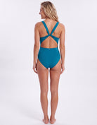 A woman modeling a teal one-piece swimsuit from the back, showing a cutout back with crisscross straps, against a plain white studio background.