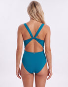 A woman viewed from the back wearing a teal one-piece swimsuit with a crossover crisscross back strap design, set against a plain white studio background.