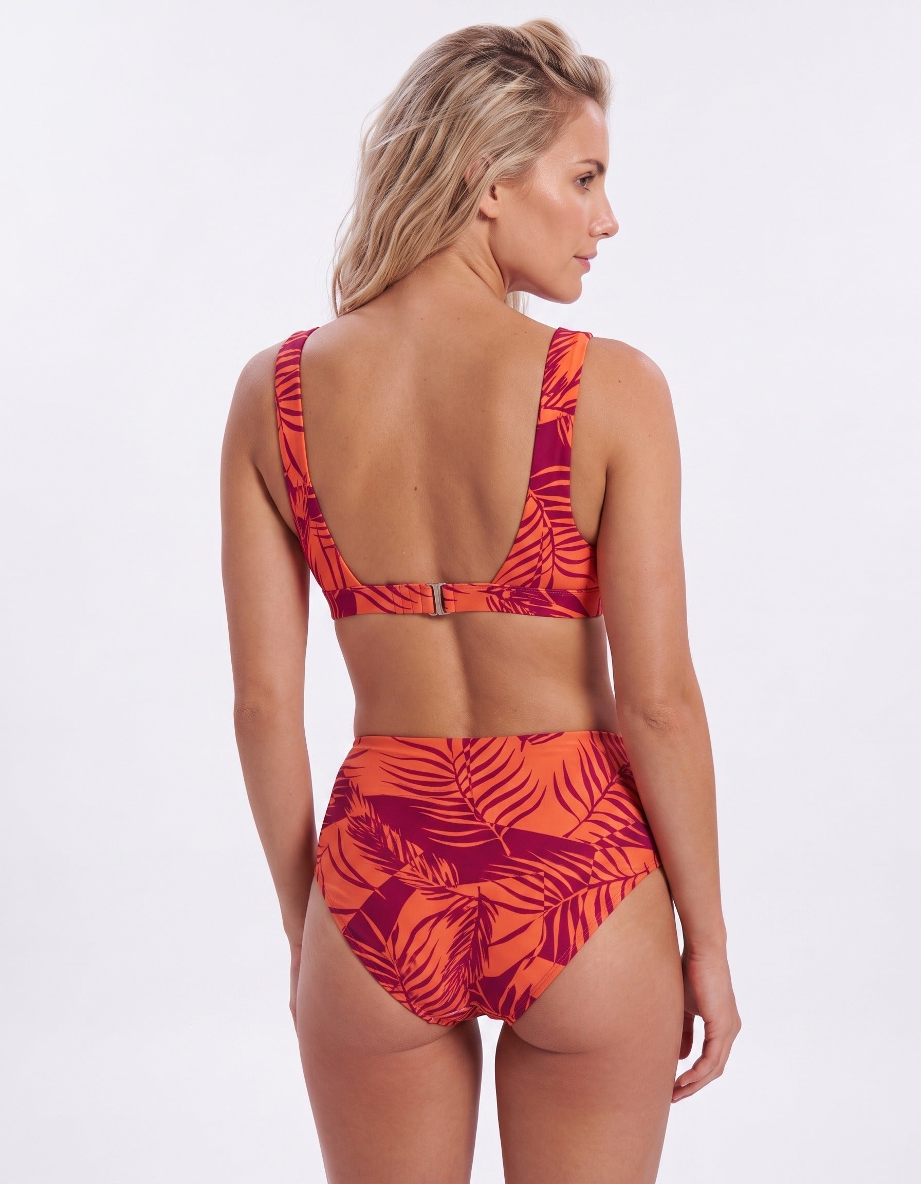 A woman modeling a two-piece bikini from the back, featuring a vibrant orange and magenta tropical leaf print against a plain white studio background.