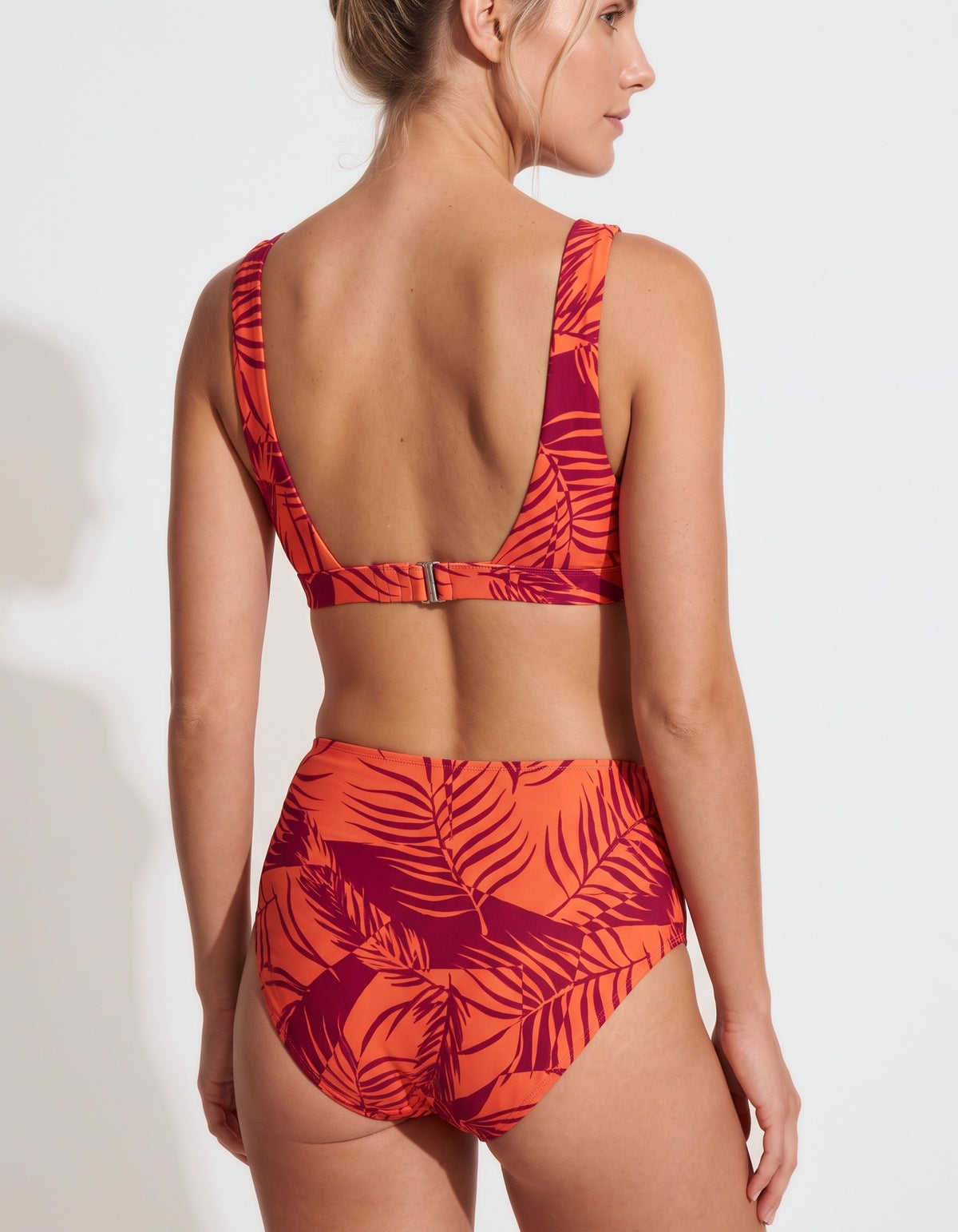 A woman shown from the back wearing a two-piece bikini with a bold tropical leaf print in bright orange and deep magenta-red. The bikini features a low open back on the top and high-waisted bottoms, photographed against a clean white studio background.