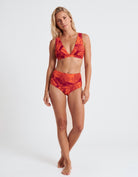 A woman modeling a two-piece bikini swimsuit with a vibrant red and orange tropical leaf pattern against a plain white studio background.