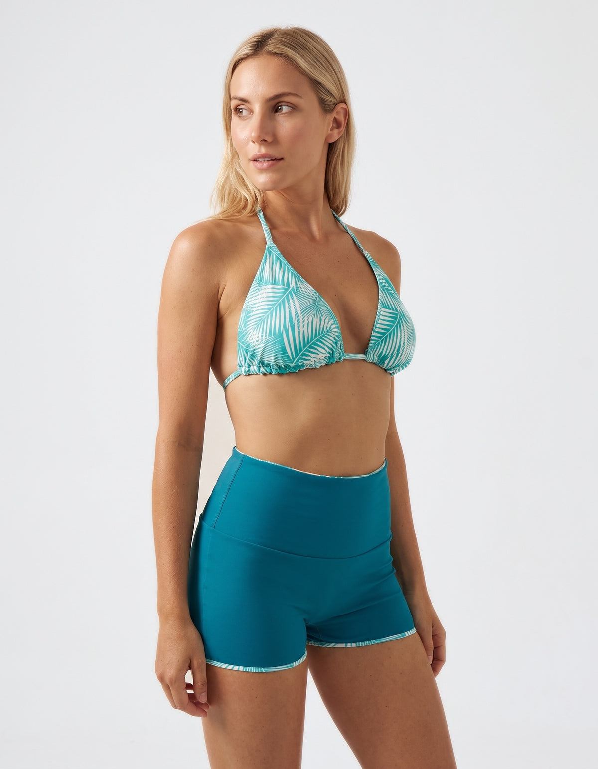 A woman modeling a two-piece swimwear set consisting of a teal and white palm-leaf patterned triangle bikini top and high-waisted teal swim shorts with a thin white trim, set against a plain light gray studio background.