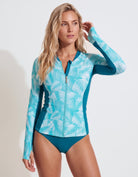 A woman modeling a long-sleeve swim rash guard and matching bikini bottoms. The rash guard features a white palm leaf pattern on a turquoise blue background with solid teal side panels and a front zipper.