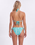 A woman shown from the back wearing a turquoise and white tropical leaf print bikini with a tie-shoulder top and side-tie bottoms, photographed against a plain white studio background.
