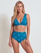 A blonde model wearing a two-piece bikini swimsuit in teal blue with a repeating hummingbird print, posed against a light gray studio background.