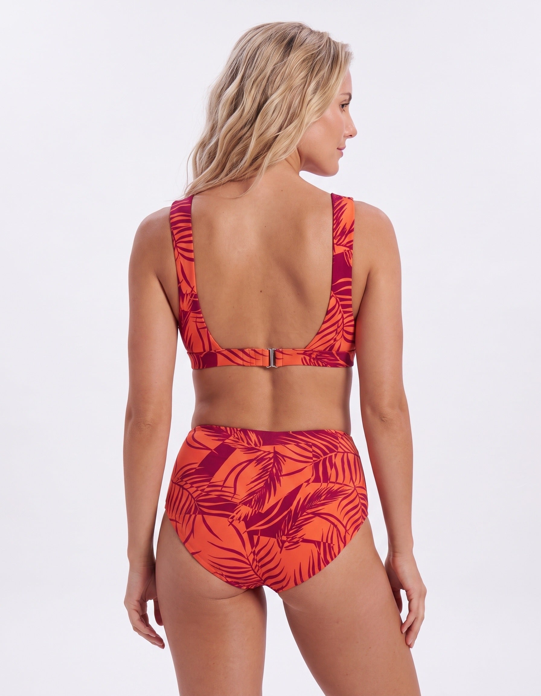 A woman shown from the back wearing a two-piece bikini with a bold tropical leaf print in orange and dark red, set against a plain white studio background.