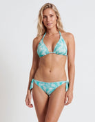 A woman modeling a two-piece bikini with a turquoise and white tropical leaf pattern against a plain white studio background.