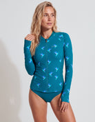 A woman modeling a teal long-sleeve full-zip swim top with a blue and purple hummingbird pattern, paired with matching teal swim bottoms, against a light gray studio background.
