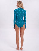 A woman stands with her back to the camera wearing a teal long-sleeve swim top with a blue bird pattern and matching teal swim bottoms against a white studio background.