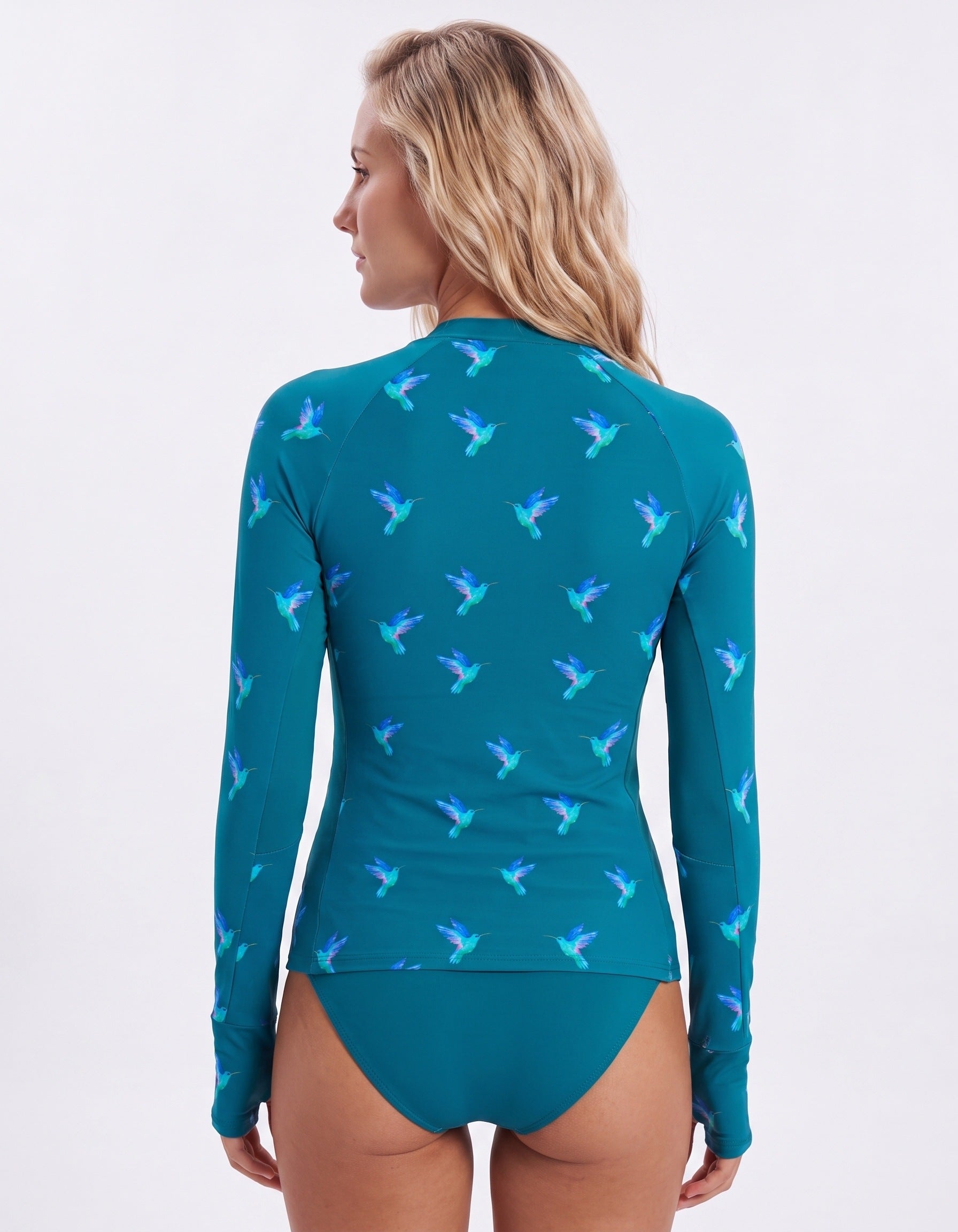 A woman shown from the back wearing a teal long-sleeve swim rash guard with a repeating hummingbird pattern in shades of blue and purple, paired with matching teal bikini bottoms, against a plain white studio background.