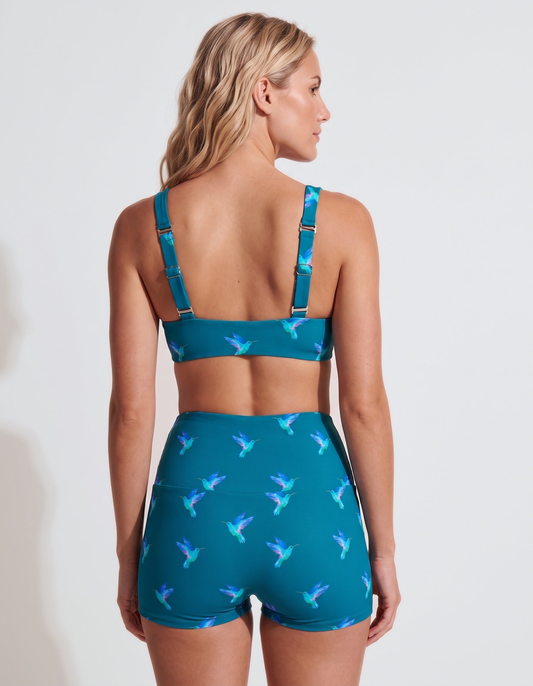 A woman shown from the back wearing a teal two-piece swimsuit with a hummingbird print, featuring a sports bra top and high-waisted shorts.
