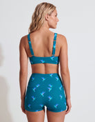 A woman shown from the back wearing a teal two-piece swimsuit with a hummingbird print, featuring a sports bra top and high-waisted shorts.