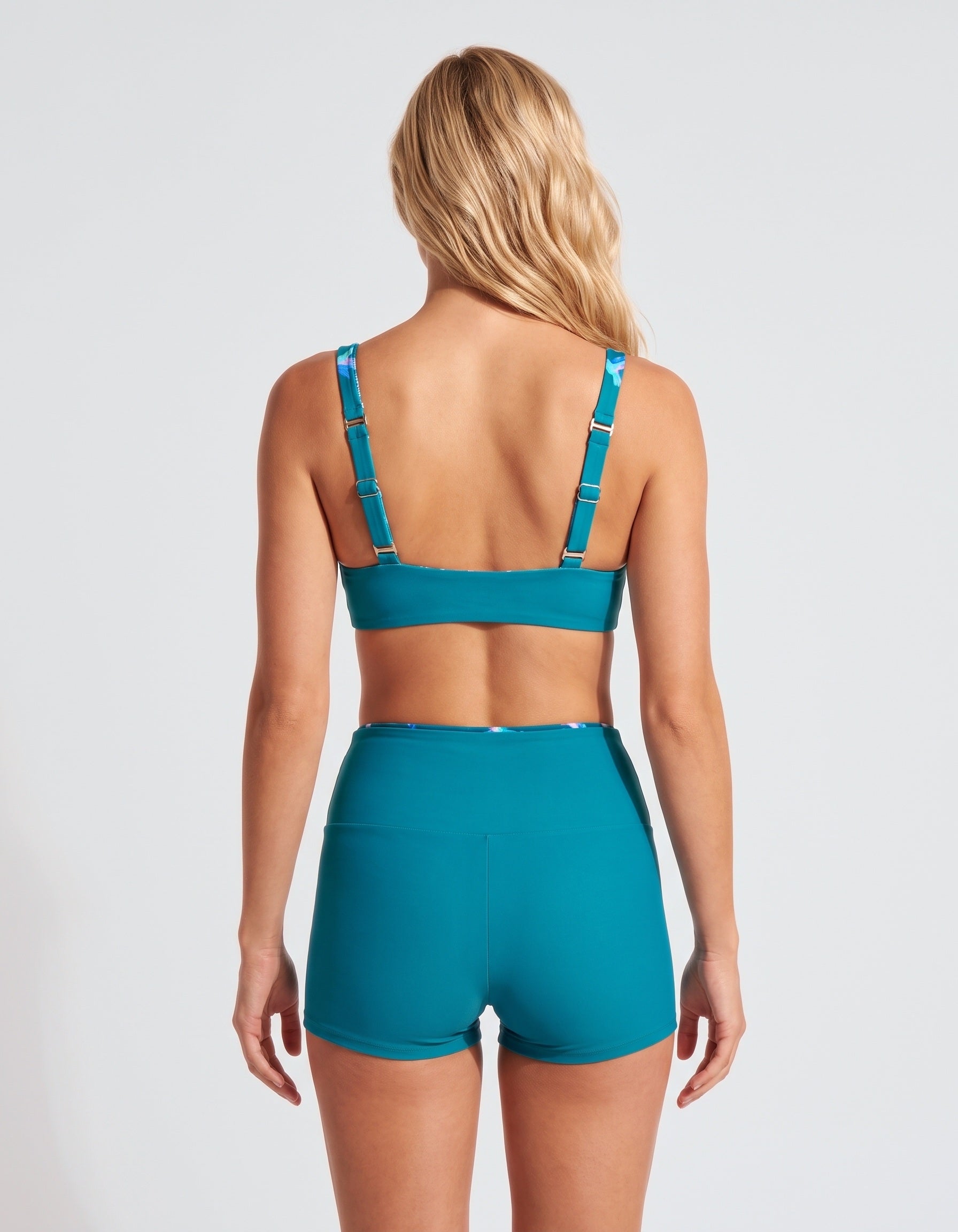 A woman shown from the back wearing a matching teal sports bra and high-waisted athletic shorts against a light gray studio background.