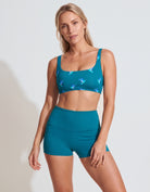 A woman modeling a teal two-piece activewear set featuring a sports bra with a blue bird pattern and matching high-waisted shorts, set against a plain light gray background.
