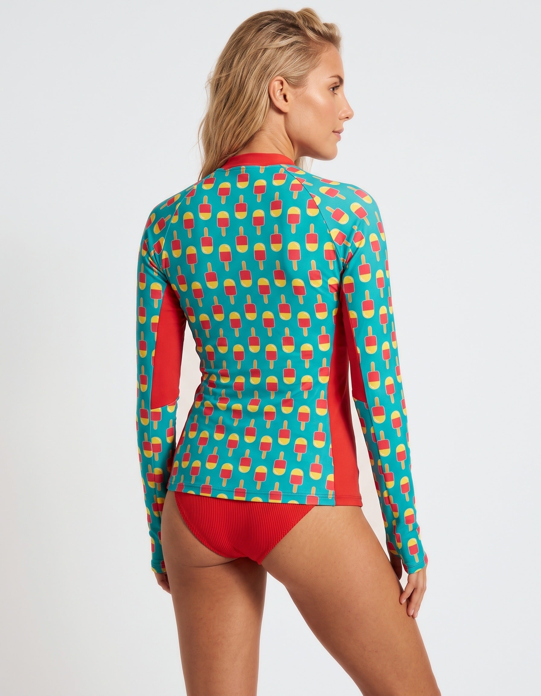 A woman modeling a long-sleeve swim top from the back, featuring a turquoise background with a repeating red and yellow ice cream cone pattern and solid red side panels and bikini bottom.