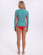 A woman stands barefoot against a plain white studio background, photographed from the back while wearing a long-sleeve swim top with a turquoise and coral geometric pattern and solid red side panels and bikini bottom.