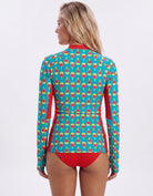 A woman shown from the back wearing a long-sleeve swim rash guard with a colorful repeating pattern on a turquoise background and solid red side panels and waistband, paired with red bikini bottoms, against a light gray studio background.