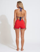 A woman stands with her back to the camera wearing a red two-piece swimsuit with blue trim and a blue neck tie, set against a plain white studio background.