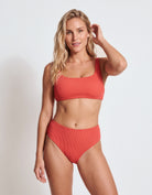 A woman modeling a ribbed two-piece bikini in a solid coral-red color, standing against a clean white studio background.
