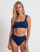 A blonde model wearing a two-piece ribbed bikini in navy blue, featuring a scoop-neck top and high-waisted bottoms, against a plain light gray studio background.