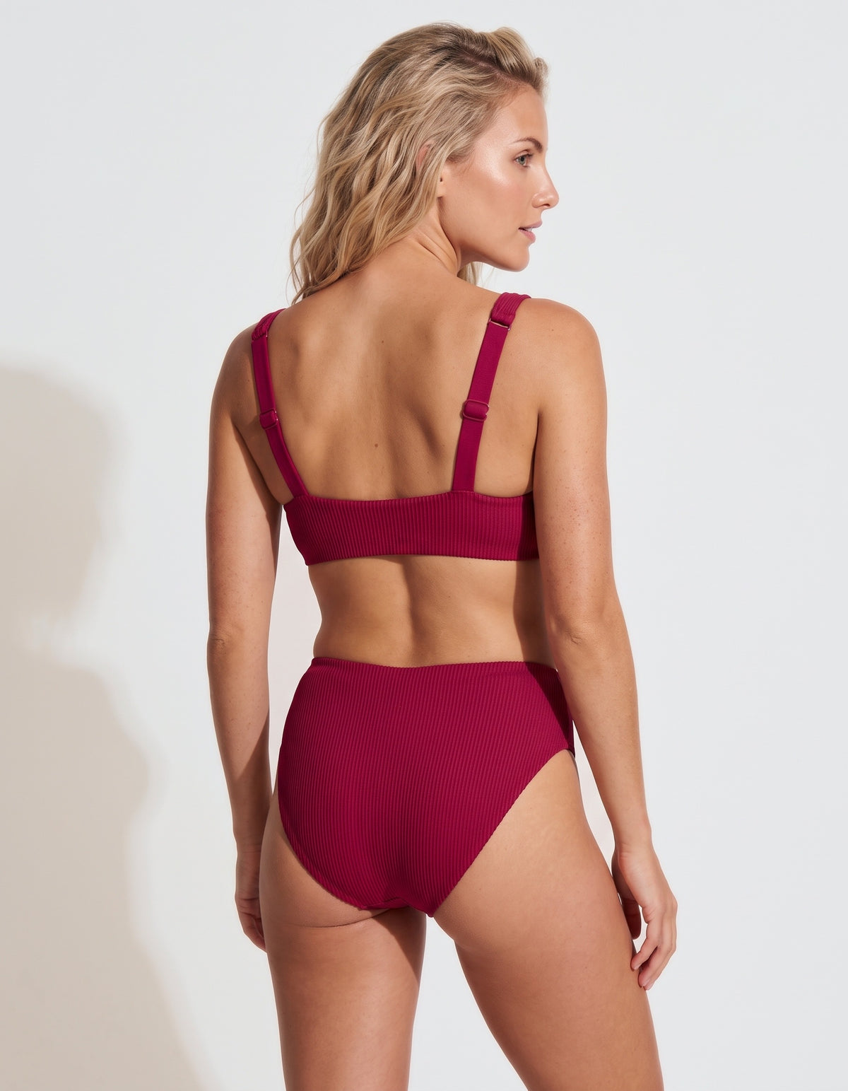 A woman modeling a ribbed two-piece swimsuit from the back, featuring a matching bikini top with wide shoulder straps and high-waisted bottoms, in a deep magenta color, set against a plain light gray background.