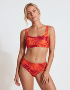 A woman modeling a two-piece bikini swimsuit with a vibrant orange and red tropical leaf print against a plain white studio background.
