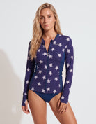 A woman modeling a long-sleeve zip-front swim rash guard in navy blue with a white sea turtle pattern, paired with matching navy swim bottoms, against a plain white studio background.