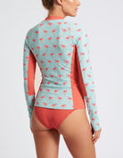 A woman shown from the back wearing a long-sleeve swim rash guard with a light blue background, pink flamingo pattern, and coral orange side panels and bottom, paired with matching ribbed coral bikini bottoms.