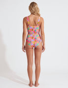 A woman stands with her back to the camera wearing a one-piece swimsuit with a vibrant tropical floral pattern in pink, blue, green, and yellow on a plain white studio background.