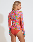A woman shown from the back wearing a long-sleeve swim top with a colorful floral print and solid coral side panels, paired with matching coral bikini bottoms, against a light gray studio background.
