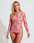 A woman modeling a long-sleeve zip-front swim top with a bright tropical floral print in pink, blue, yellow, and green, paired with red bikini bottoms, against a clean white studio background.
