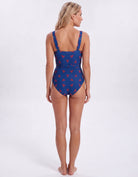 A woman modeling a one-piece swimsuit from the back, featuring a royal blue base with a red and white tropical floral pattern, set against a plain white studio background.