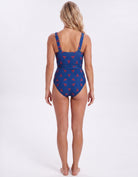 A woman viewed from the back wearing a one-piece swimsuit with a blue base and a red pattern, standing against a plain white studio background.