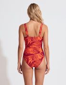 A woman viewed from the back wearing a one-piece swimsuit with a red and orange tropical leaf pattern against a light gray studio background.