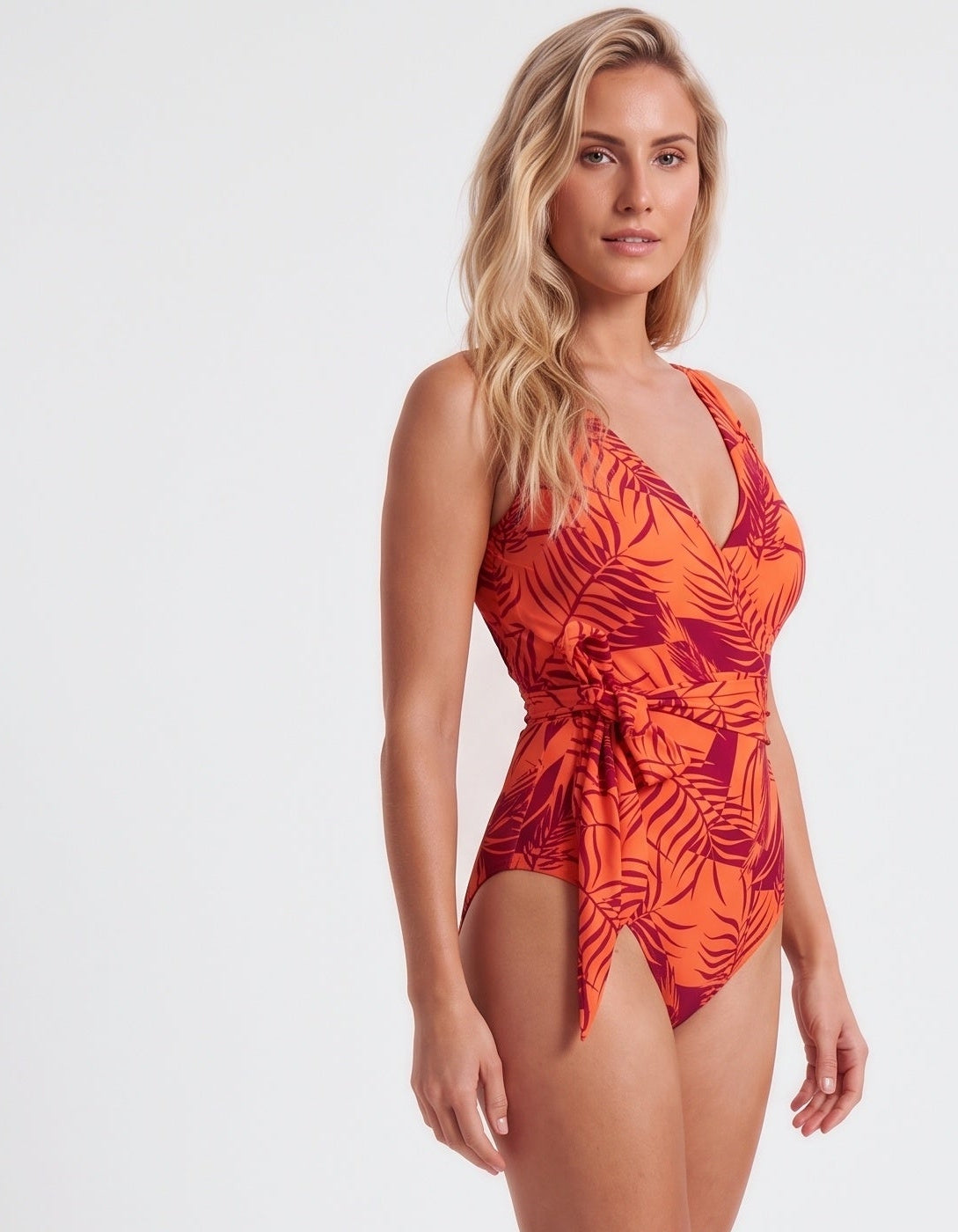 A woman modeling a one-piece swimsuit with a vibrant orange and red tropical leaf pattern against a plain white studio background.