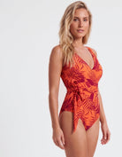 A woman modeling a one-piece swimsuit with a vibrant orange and red tropical leaf pattern against a plain white studio background.