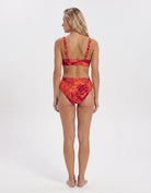 A woman stands with her back to the camera wearing a two-piece bikini with a red and orange tropical leaf pattern on a white studio background.
