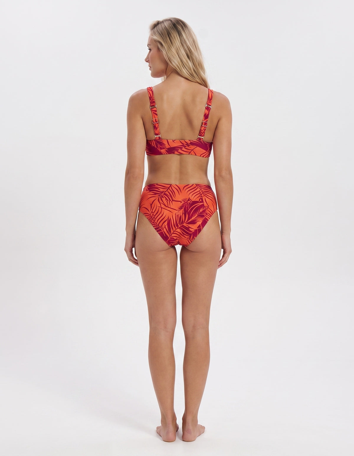 A woman stands with her back to the camera wearing a two-piece bikini with a red and orange tropical leaf pattern on a white studio background.
