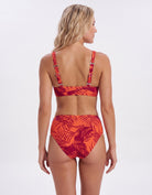 A woman shown from the back wearing a two-piece bikini with a red and orange tropical leaf print, set against a plain white studio background.
