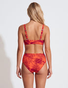 A woman shown from the back wearing a two-piece bikini with a red and orange tropical leaf print, set against a plain light gray studio background.